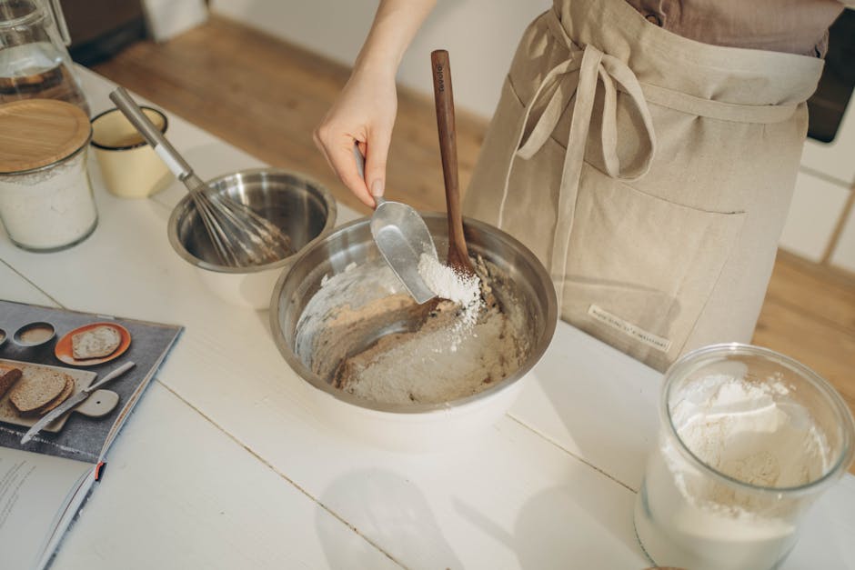 Person adding flour to dough in a stainless steel bowl while baking at home.